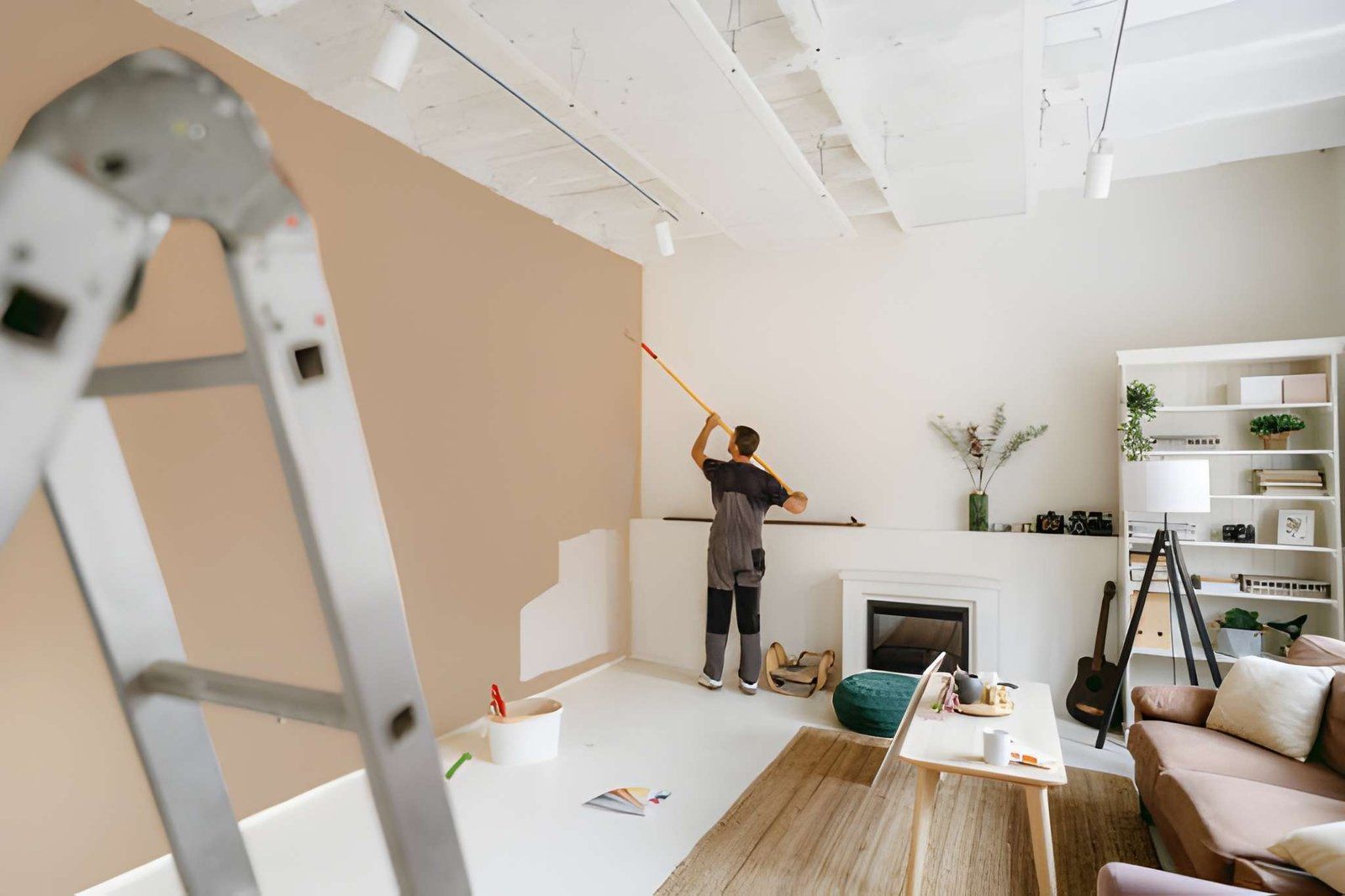 Person painting a tan wall in a bright living room with a ladder in the foreground
