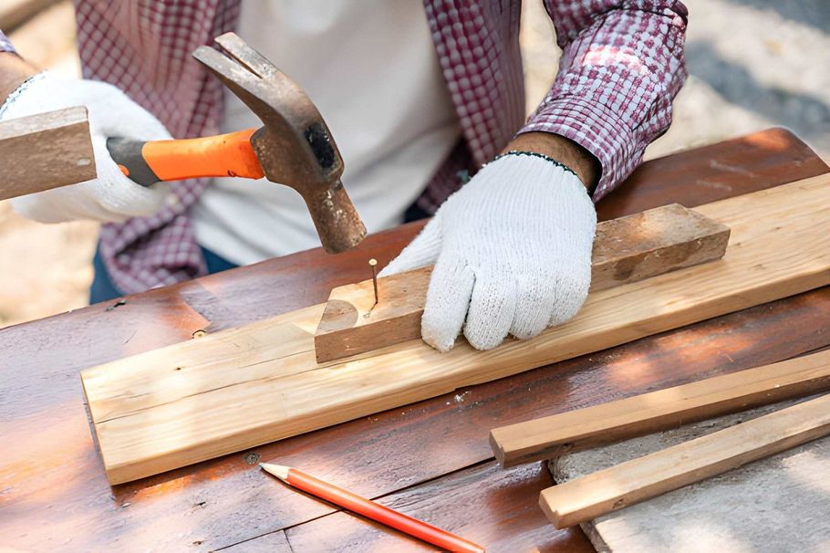 Gloved hands hammer a chisel into a wooden board on a workbench with tools nearby