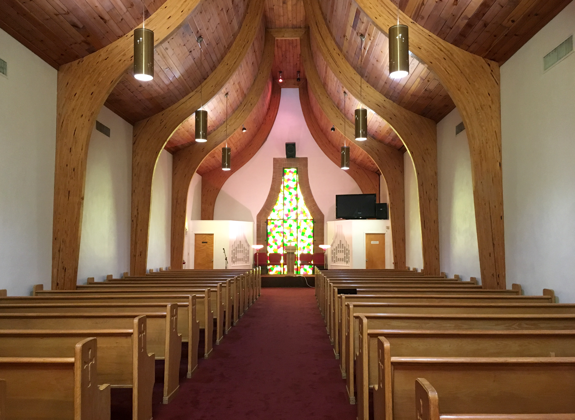 Interior of a church with wooden arched beams, rows of wooden pews on both sides of a central aisle, and a stained glass.