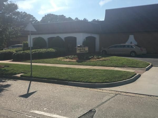 A single-story commercial building with a white front facade, green landscaping, a sign, and a car in the parking lot.