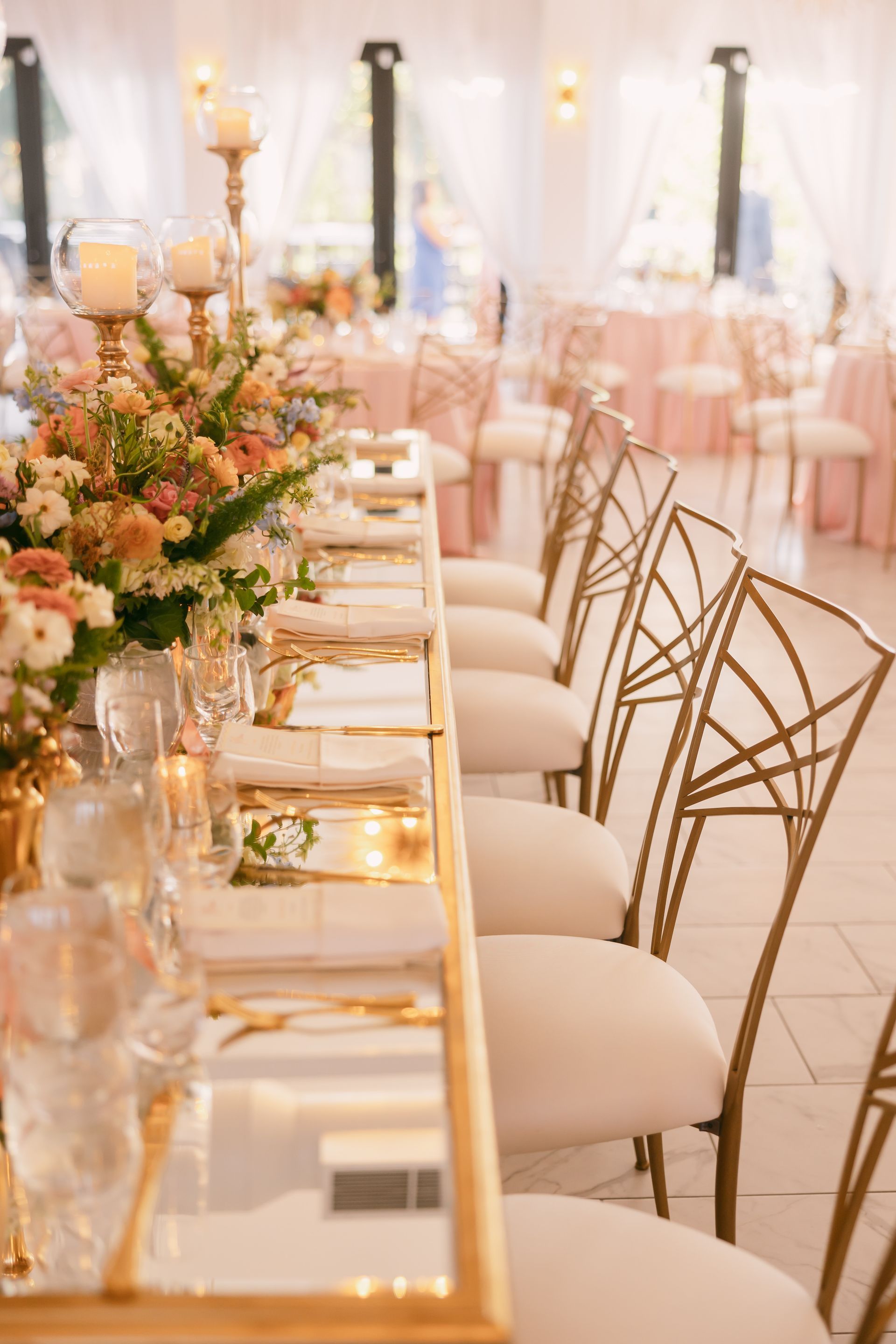 Elegant wedding reception table set with flowers, candles, and gold chairs in a bright, formal indoor setting.