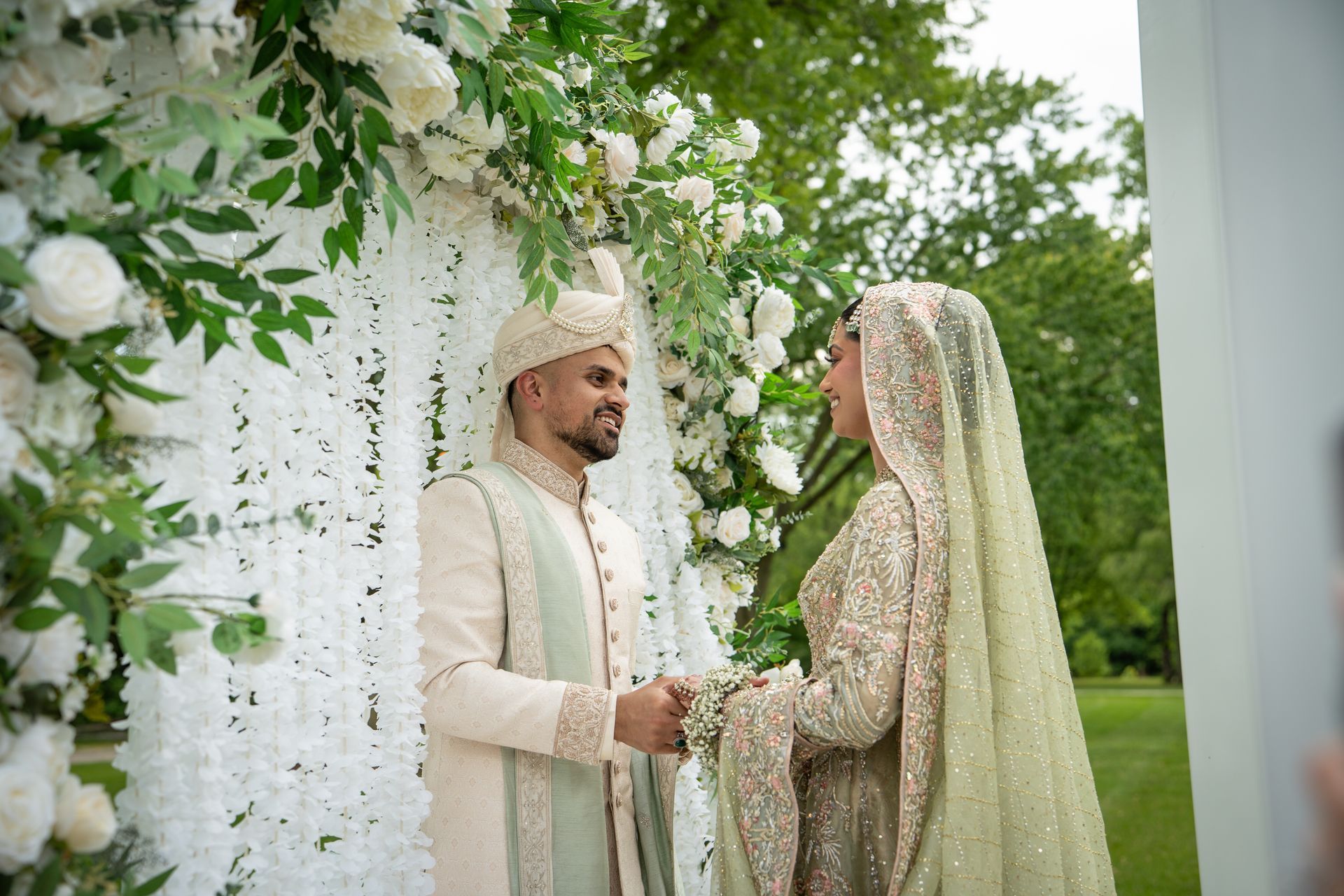 A couple in traditional wedding attire holding hands while facing each other under a flower-adorned arch outdoors.