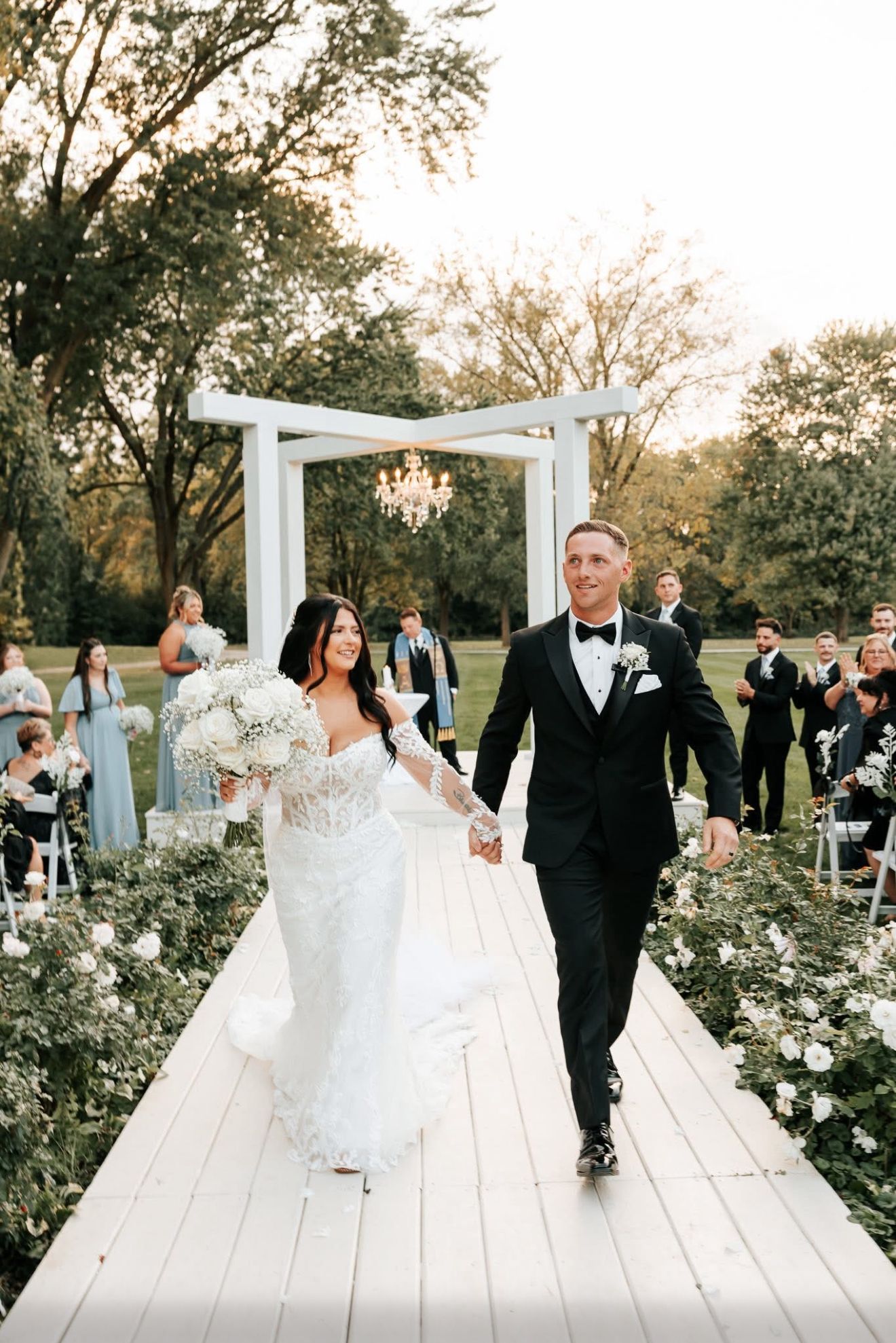 A bride and groom walk hand-in-hand down a wooden aisle outdoors, smiling after their wedding ceremony.
