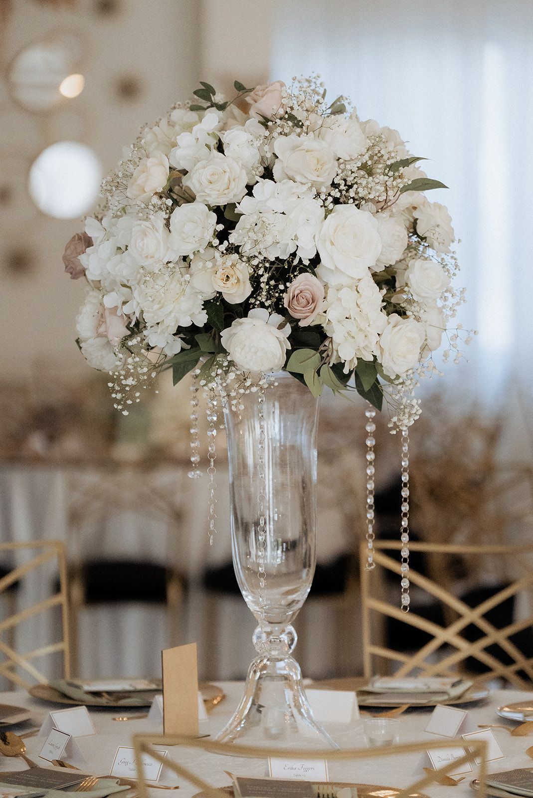A large, elegant white floral centerpiece with hanging crystals in a tall glass vase on a decorated event table.