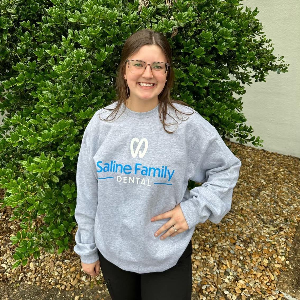 A woman wearing a saline family dental sweatshirt is standing in front of a bush.