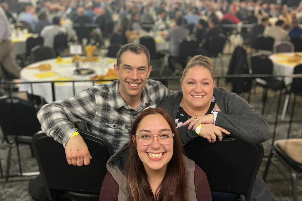 Three people smiling, seated in front of a crowd at an event, with tables in the background.