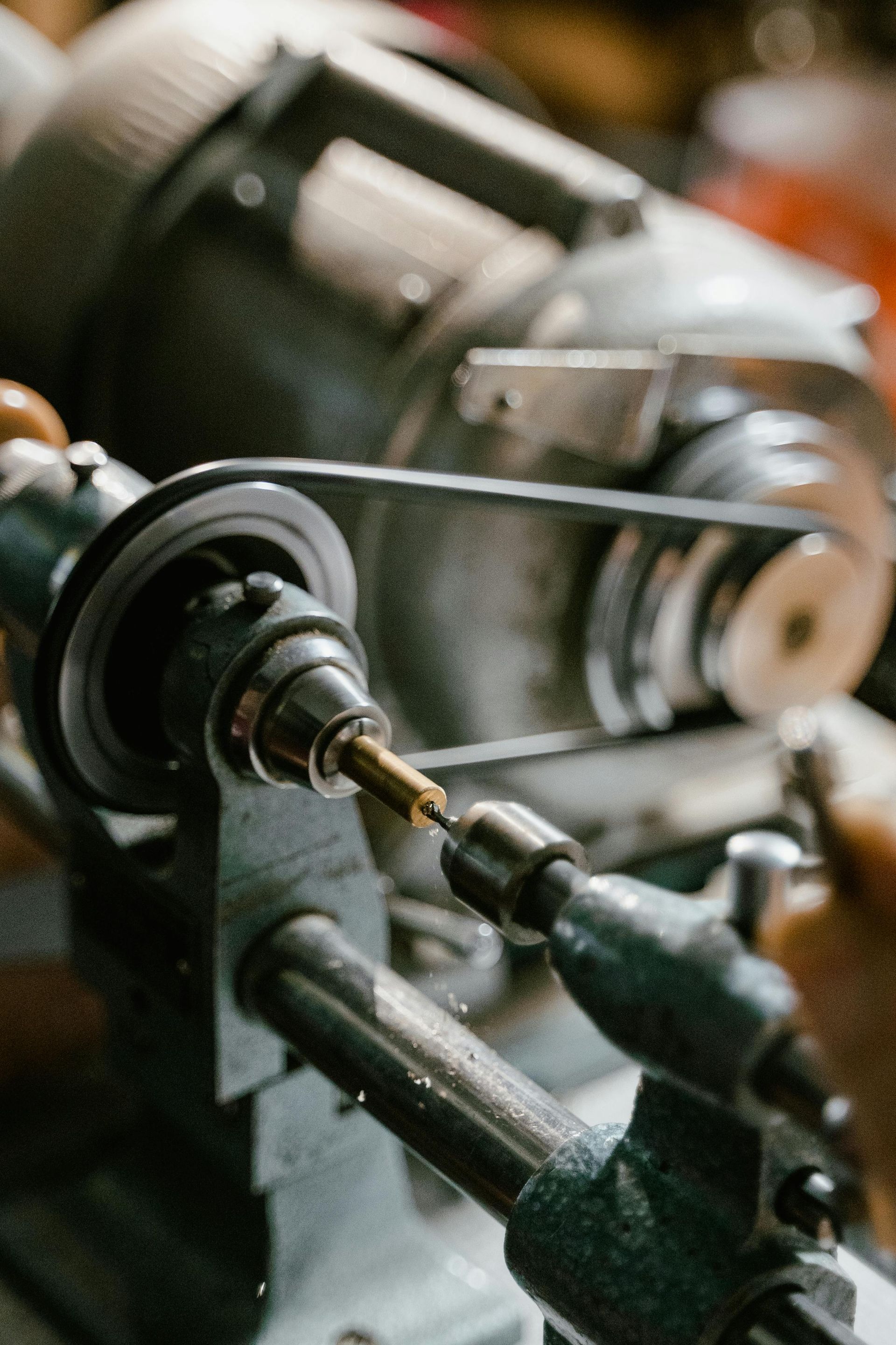 A metal lathe in use, focused on a turning gold-colored piece. A belt drives the rotating mechanism.