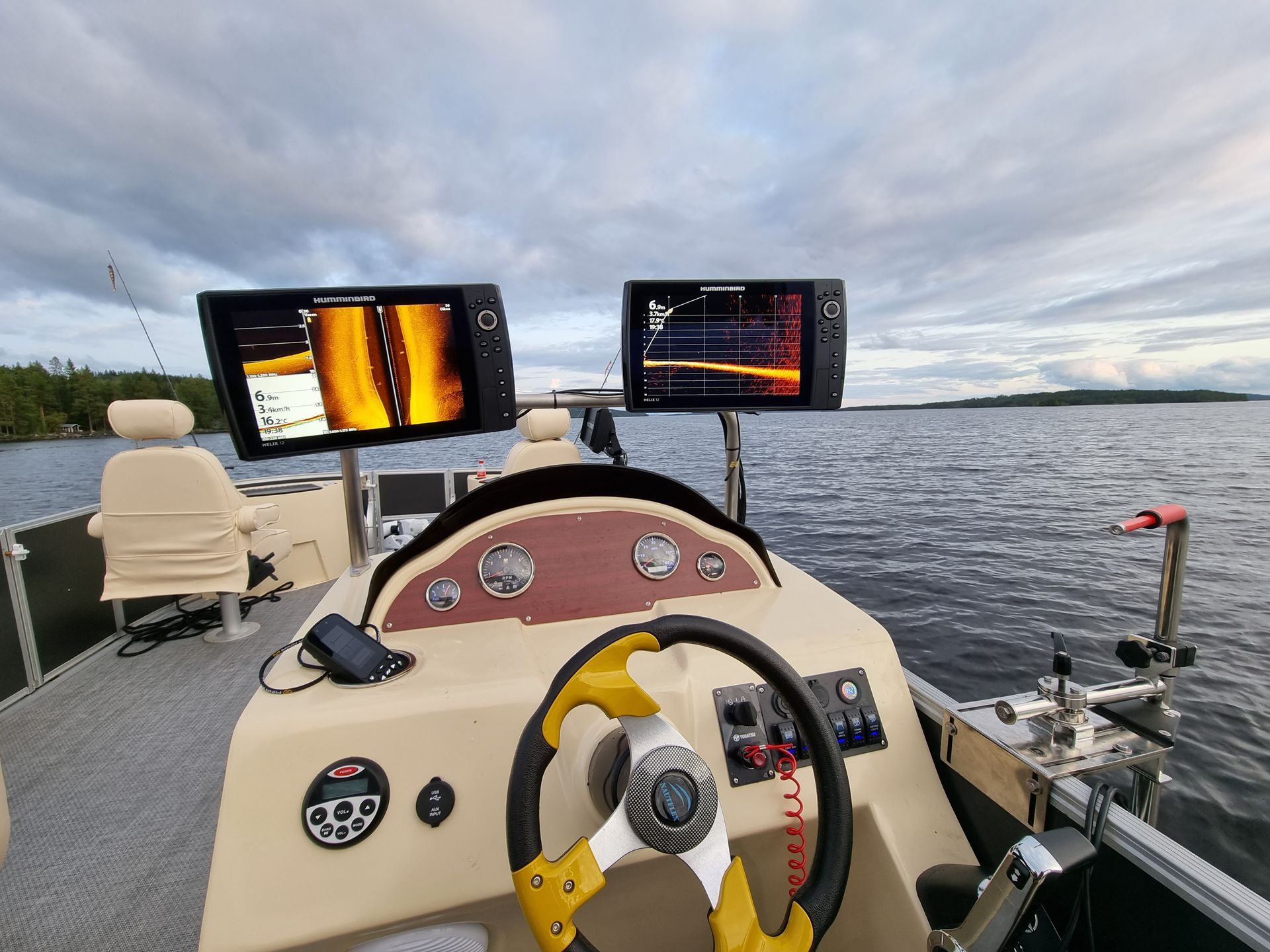 Boat helm with steering wheel and twin fish finder screens on a calm body of water under cloudy skies
