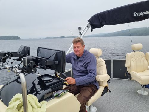 Man seated at a boat helm with fishing gear on a calm lake under an overcast sky