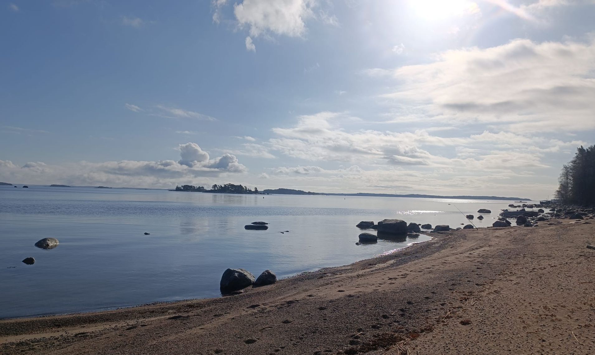 Calm rocky shoreline under a bright sky with clouds and distant islands on the horizon
