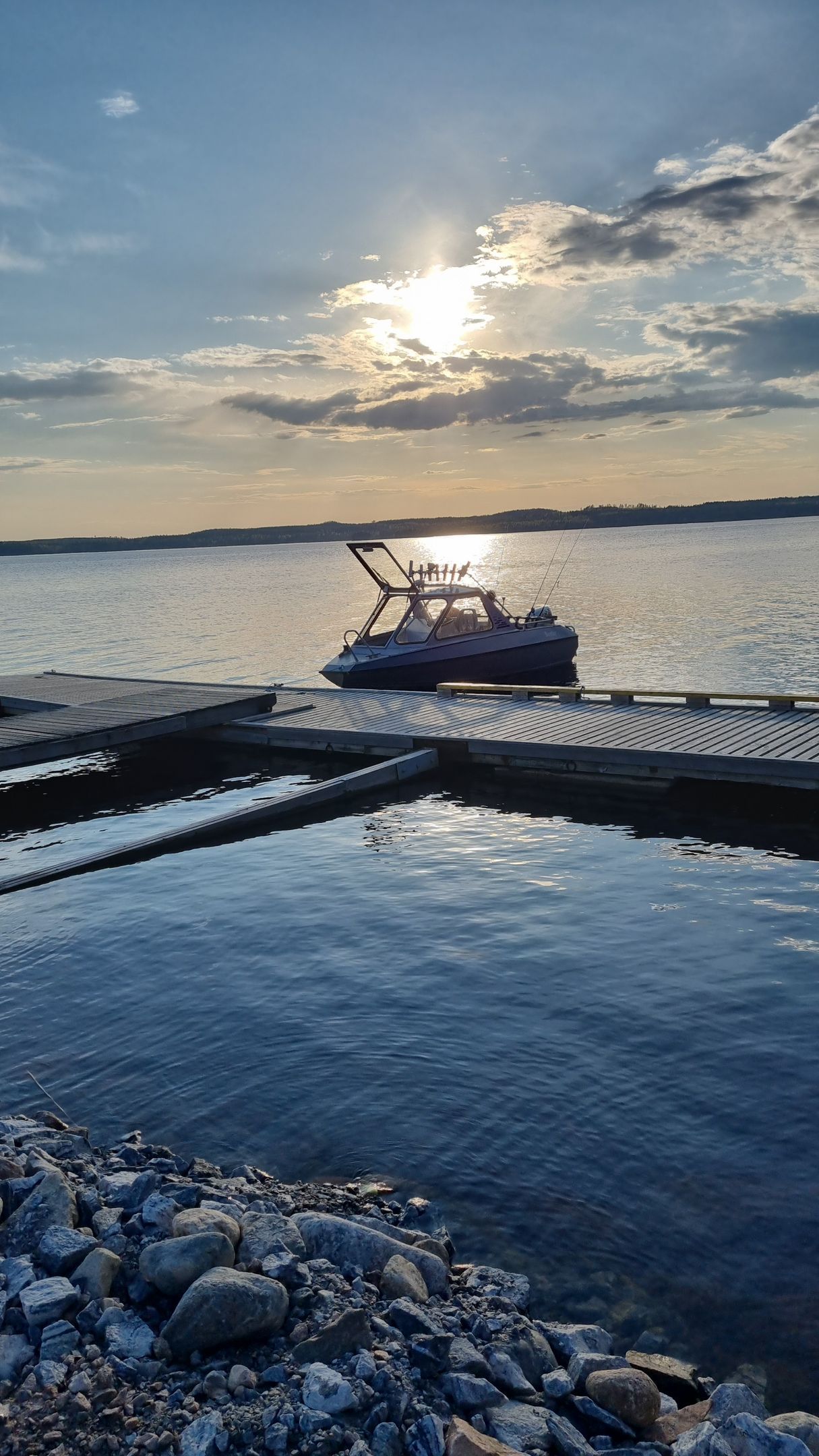 Boat docked at a lake harbor at sunset, with calm water and rocky shoreline in the foreground.