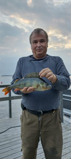 Man holding a perch on a boat at sunset with calm water and cloudy sky.