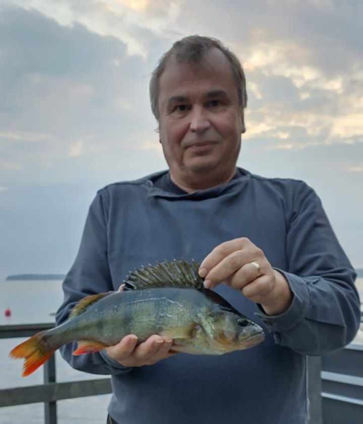 Man holding a perch on a dock at dusk, with calm water and cloudy sky behind him