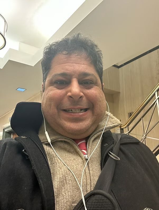 Smiling person in a dark coat with earphones, standing indoors near a staircase and railings.