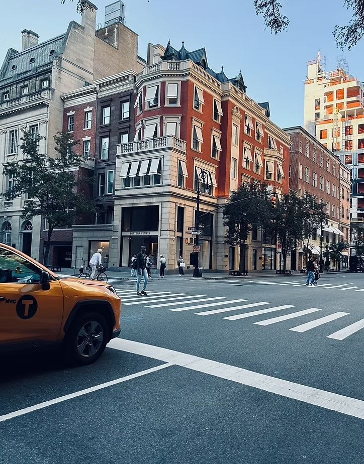 Urban street corner with red brick buildings, pedestrians, and a yellow taxi at a crosswalk.