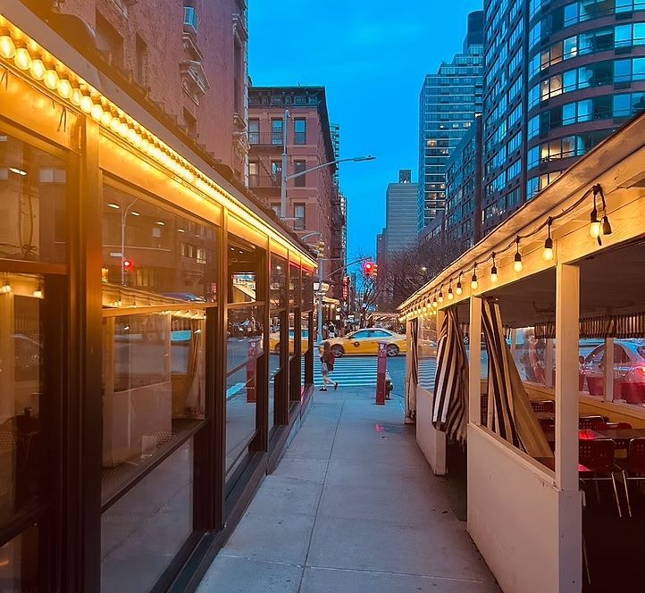City sidewalk between lit restaurant patios at dusk, with modern buildings in the background.