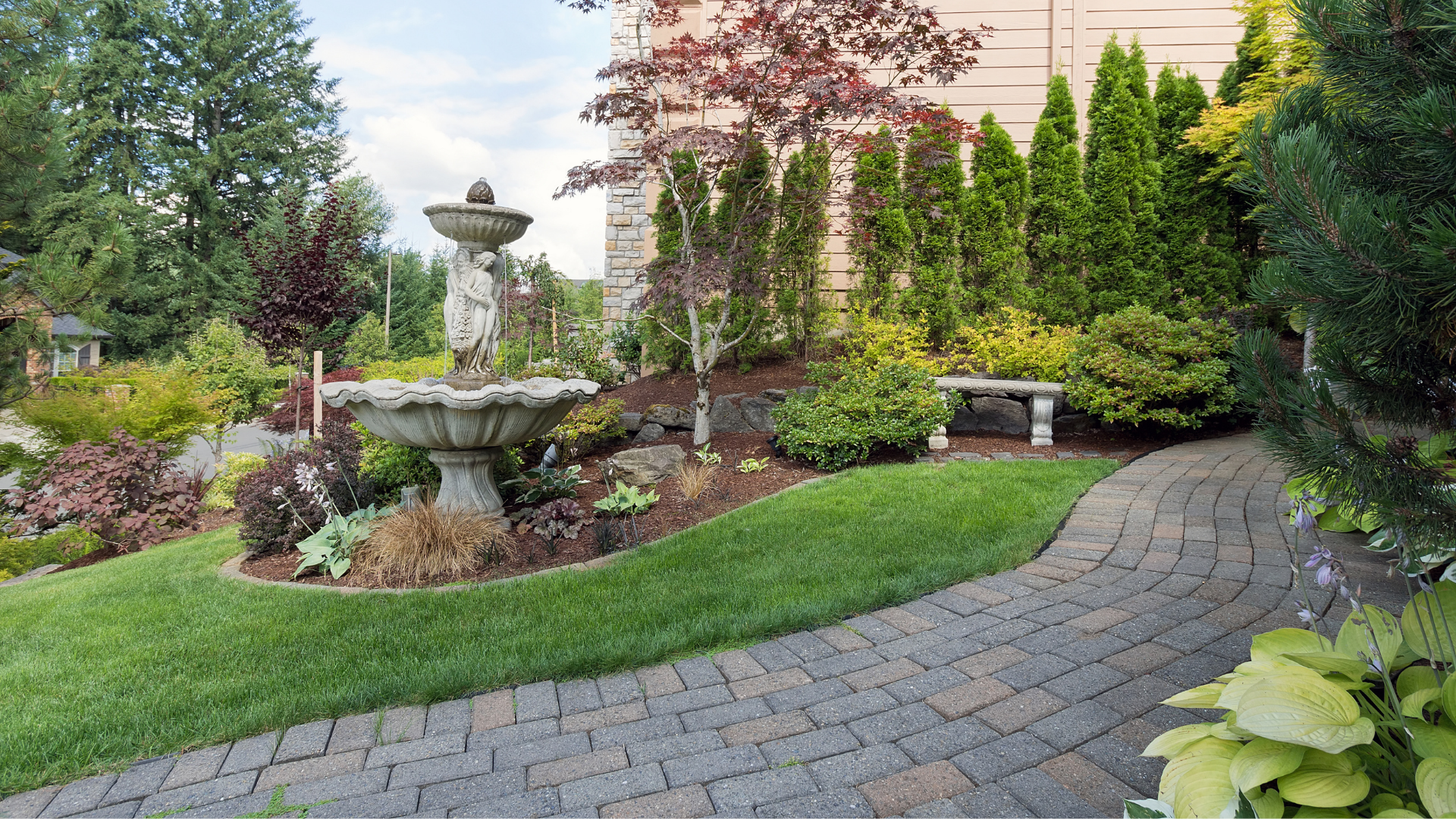 Landscaped garden with stone fountain, curved paver path, benches, and lush greenery