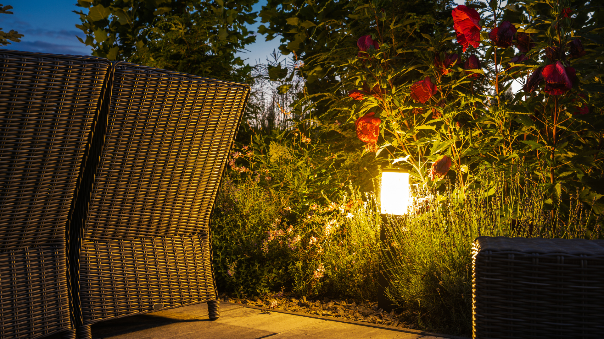 Outdoor garden seating with wicker chairs, warm lamp light, and red flowers at dusk