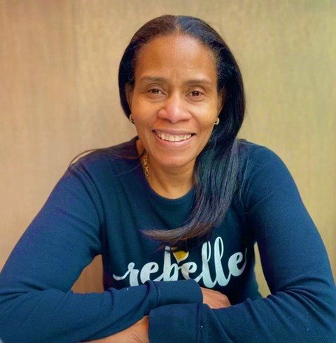 Smiling person in a navy shirt leaning on folded arms against a beige background.