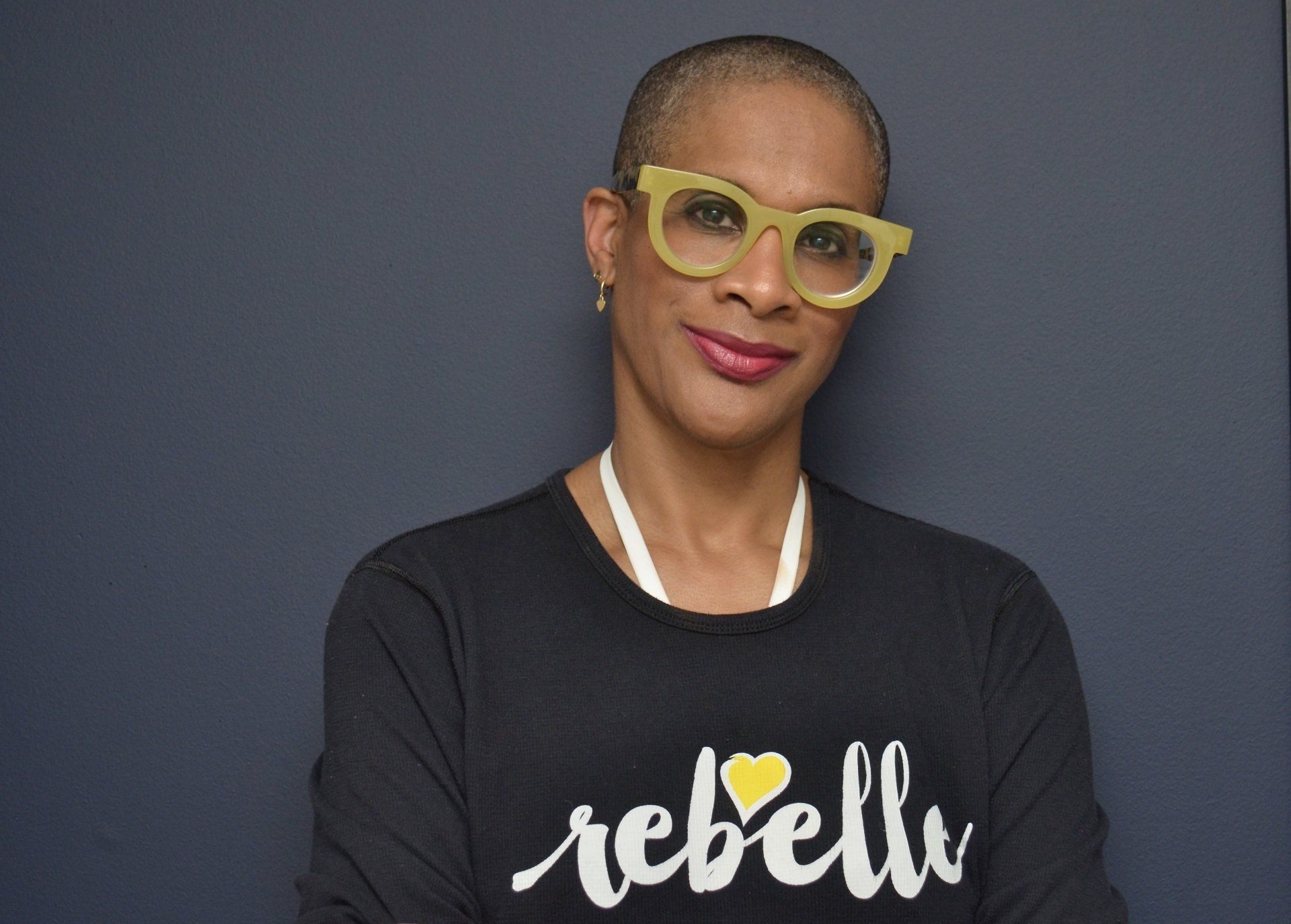 Person wearing yellow glasses and a black “rebel” shirt against a gray background