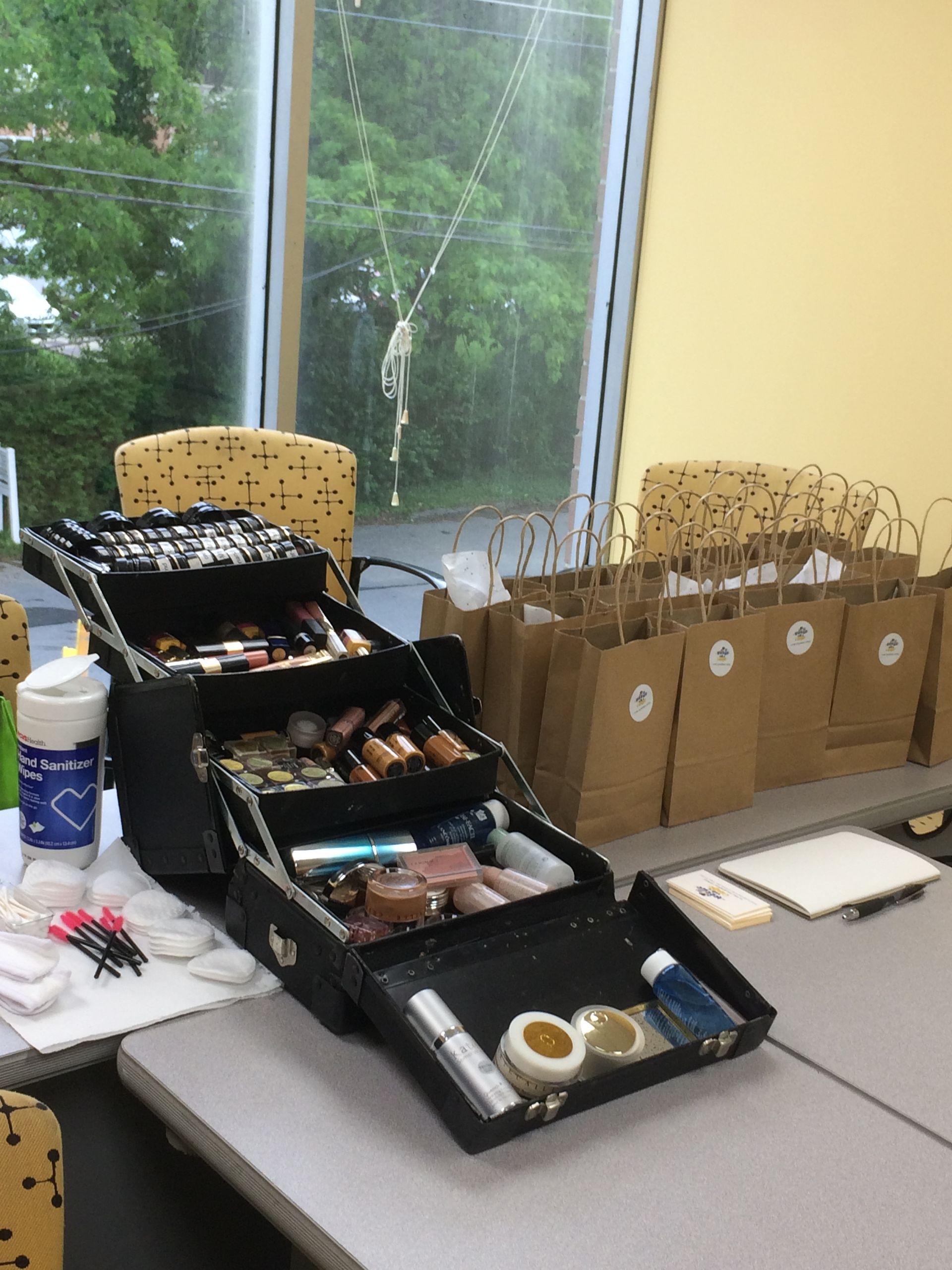 Makeup kit and paper bags on a table by a window, with green trees outside