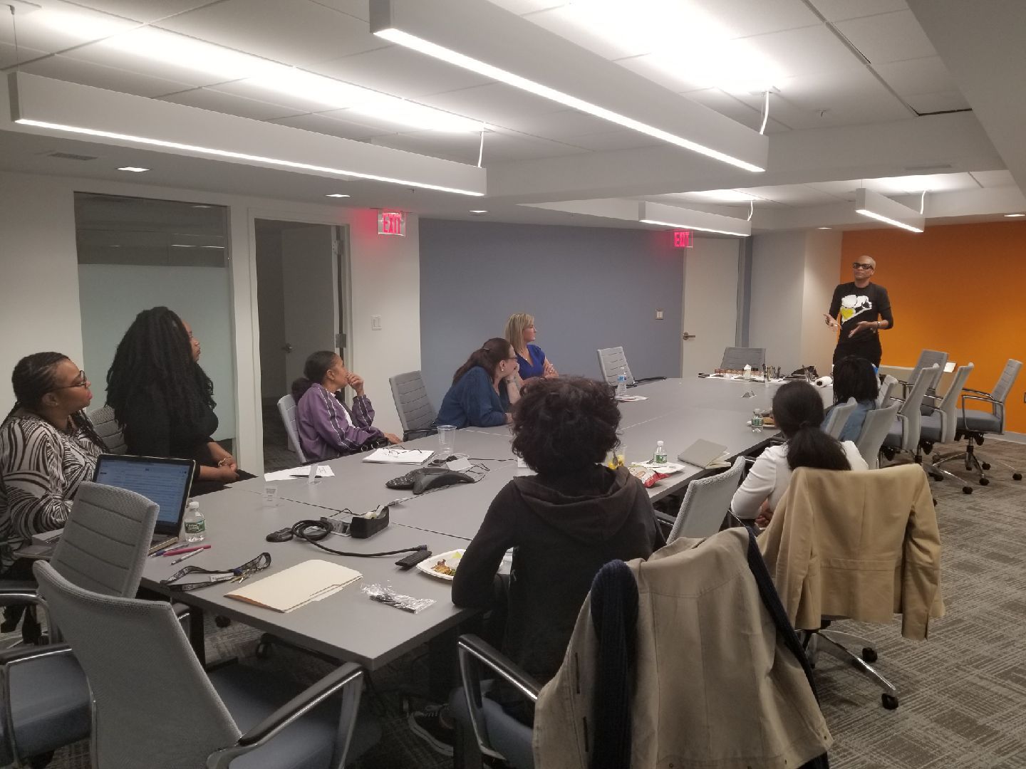 People seated around a conference table in a meeting room, listening to a speaker at the front.
