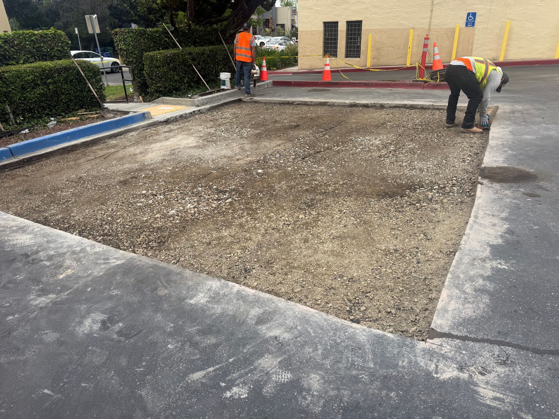 Workers leveling a gravel area beside a building, with orange safety gear and barriers nearby