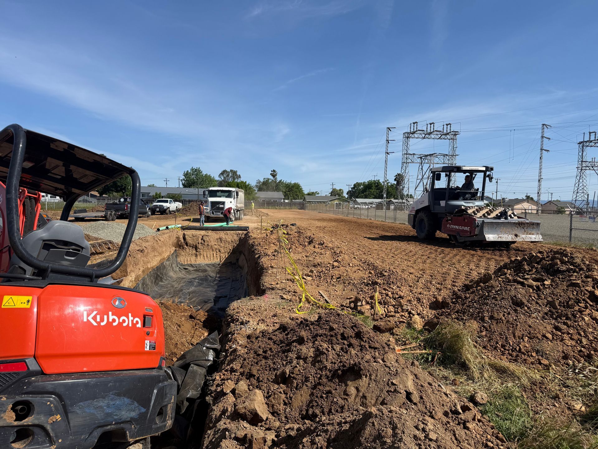 Construction vehicles on a dirt road, with a red Kubota machine in the foreground and a skid steer in the distance.