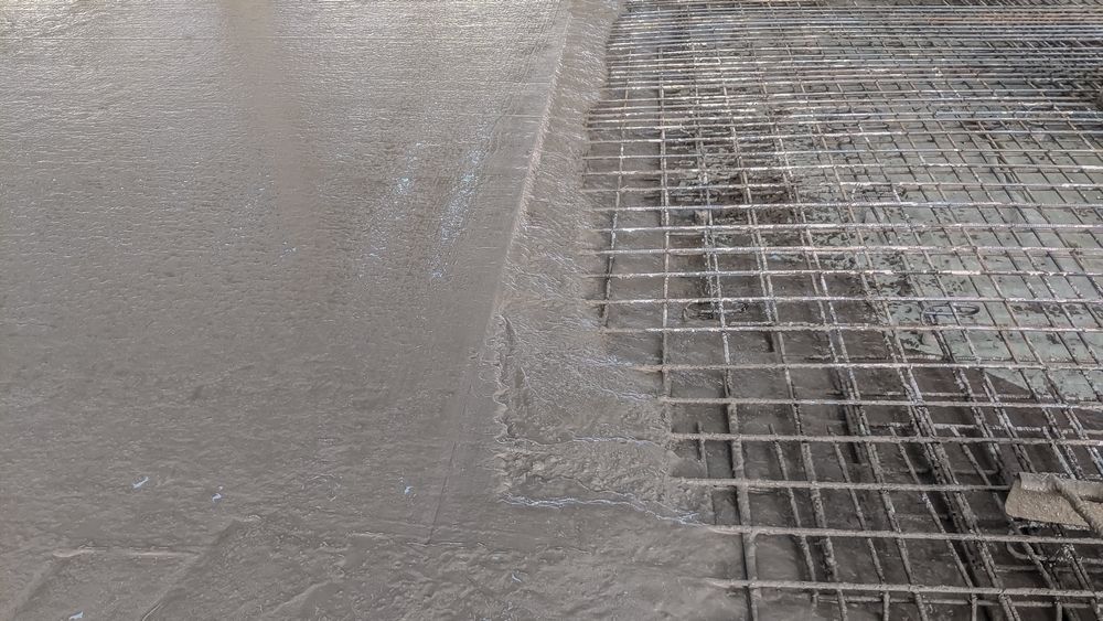Wet concrete being smoothed beside a metal rebar grid at a construction site
