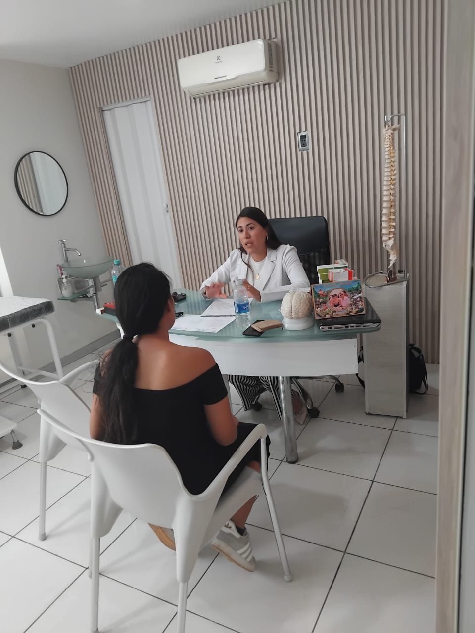 Mujer con bata blanca hablando con una persona sentada en una oficina.