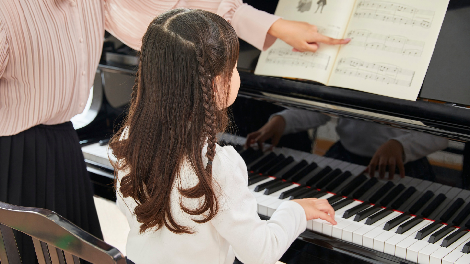 Child playing piano while an adult points to the sheet music nearby