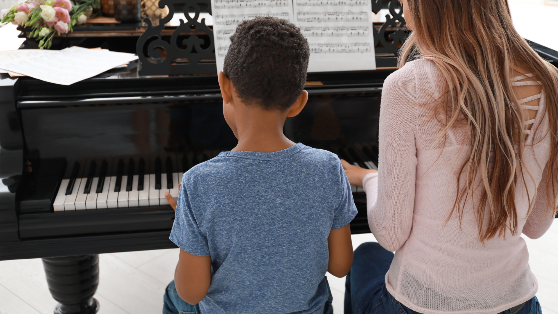 Two children sit at a piano, one playing while sheet music rests on the stand.