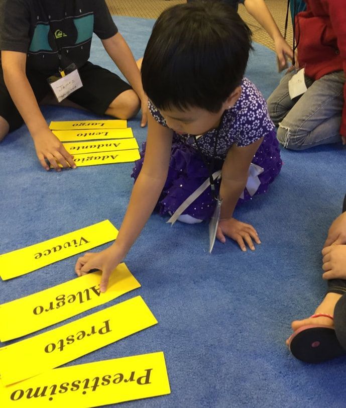 Child in purple dress reaching for yellow word cards on a blue carpet during a group activity