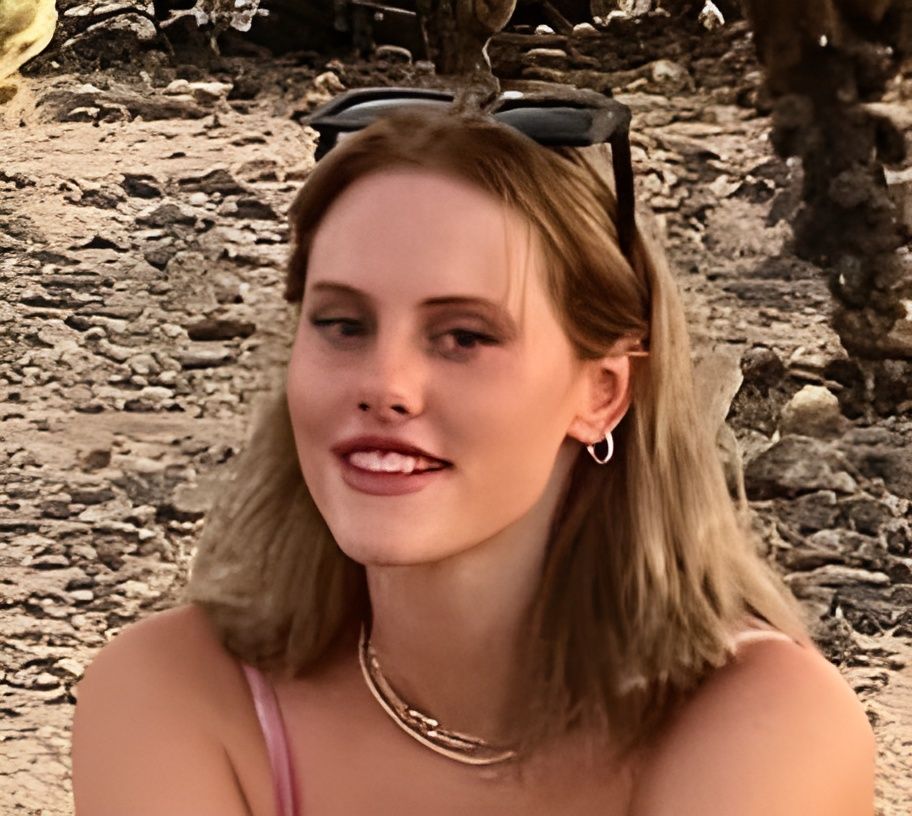 Woman smiling on a rocky beach, wearing sunglasses on her head and a pink top.