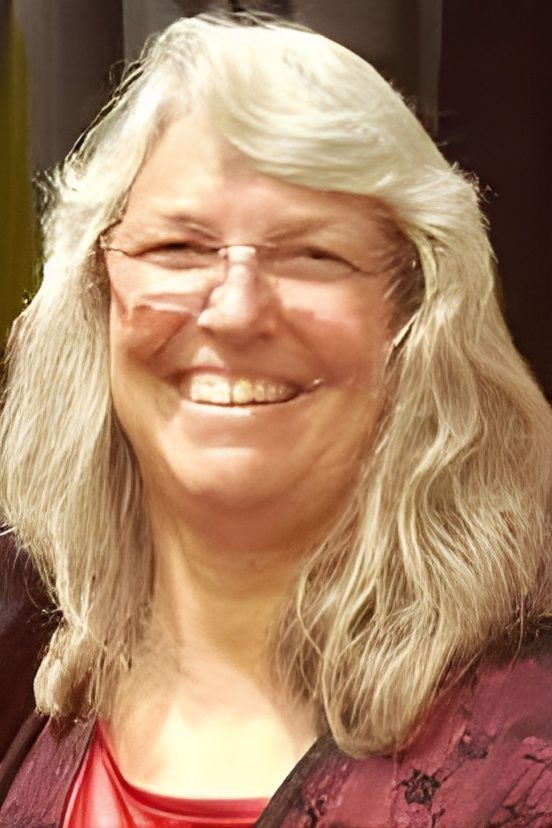Smiling woman with long gray hair and glasses, wearing a maroon top against a dark background