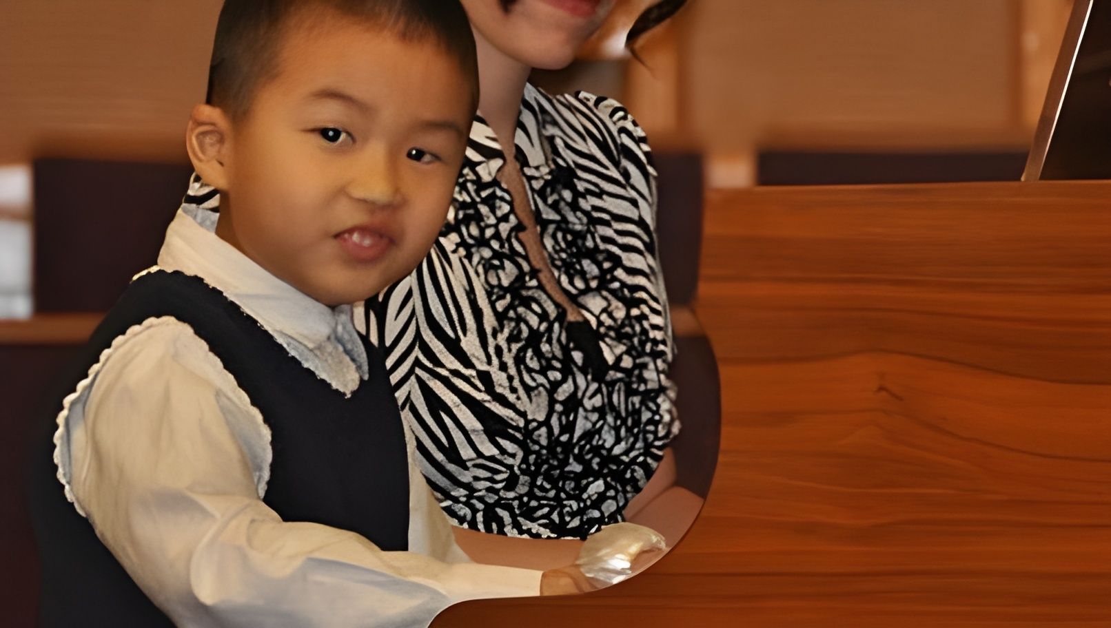 Child in a white shirt and navy vest sitting beside an adult at a wooden piano, both indoors.