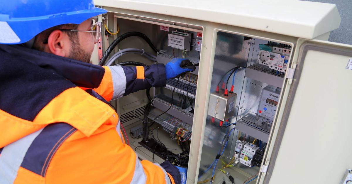 Técnico con chaleco de seguridad naranja y casco azul trabajando dentro de un armario de control eléctrico abierto.