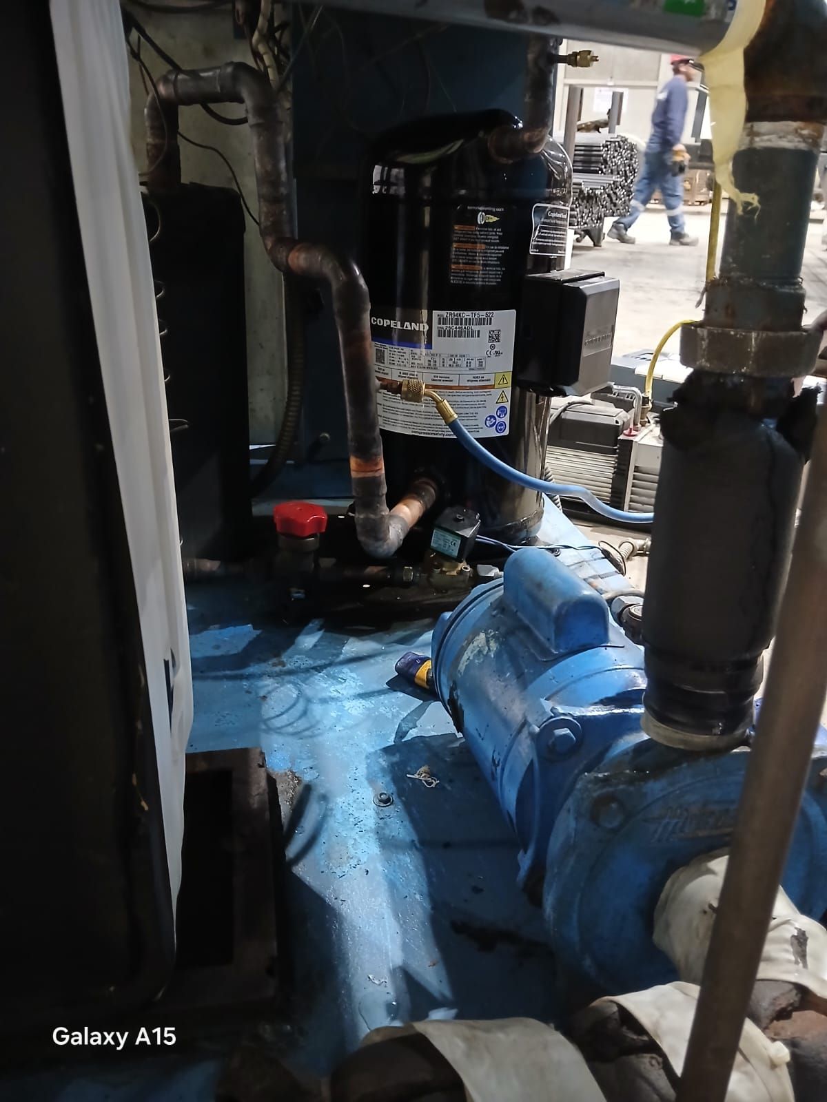 Bomba industrial azul y tuberías en una sala de máquinas con poca luz, con una persona visible al fondo.