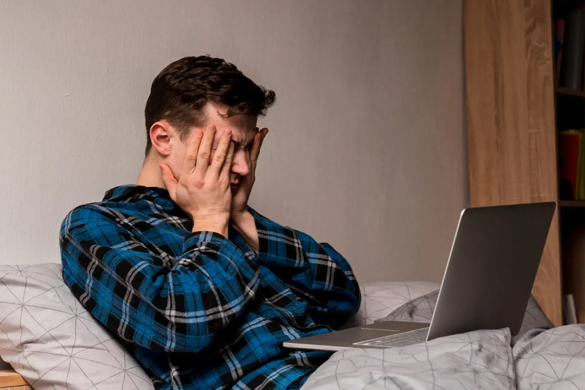 Man covering face while sitting in bed beside an open laptop, looking stressed.