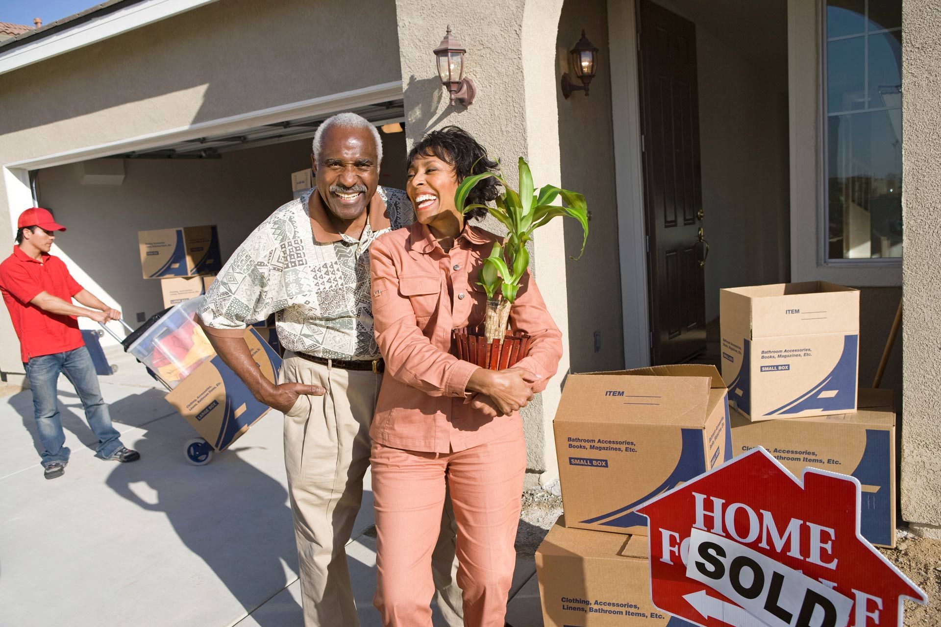 Couple in front of new home, holding plant, with moving boxes and 