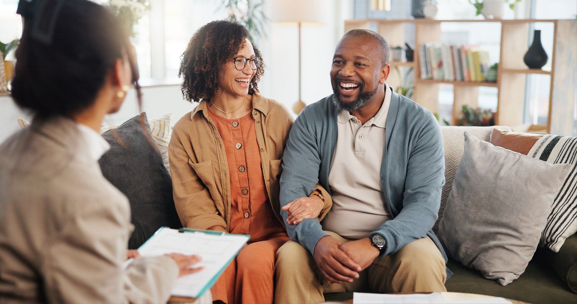 Couple smiles at a person holding a clipboard on a couch; indoors.