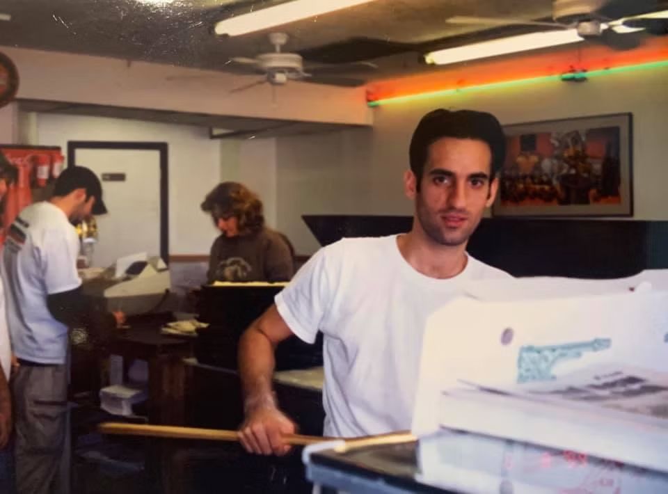 Man in white t-shirt holds pizza peel at counter, other workers behind him in a pizza restaurant.