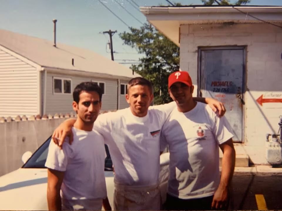 Three men with arms around each other pose outside a building; one wears a Phillies hat.