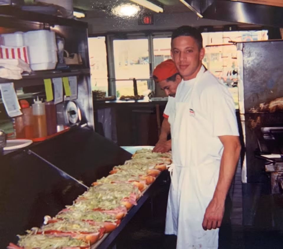 Man in white apron smiles near a long sub sandwich being prepared in a commercial kitchen.