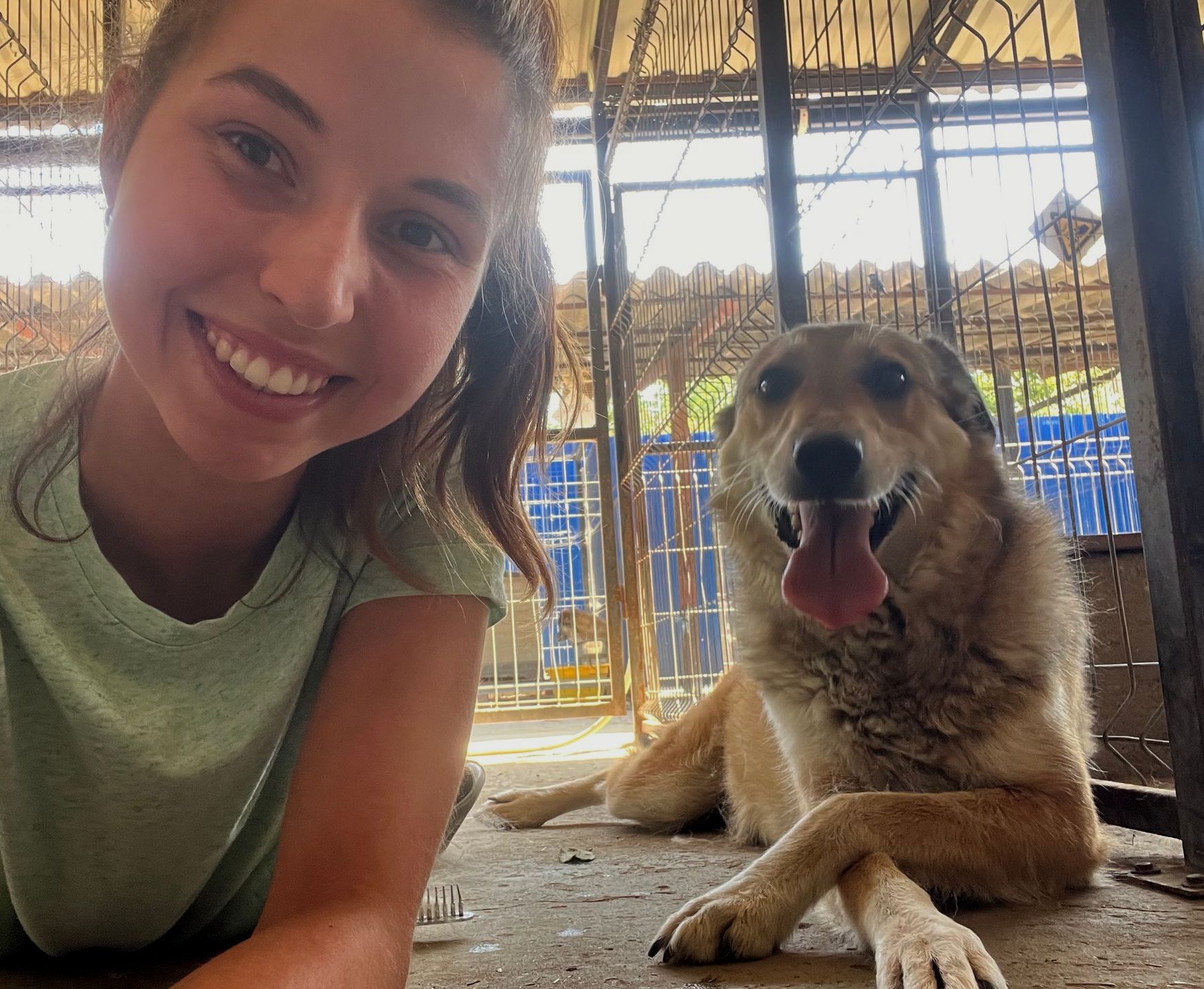 Een lachende vrouw leunt naast een blije hond die op de grond ligt onder een afdak.