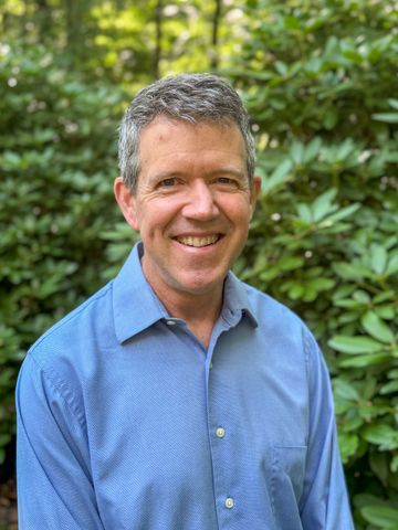 Smiling man in a blue shirt standing outdoors in front of green foliage