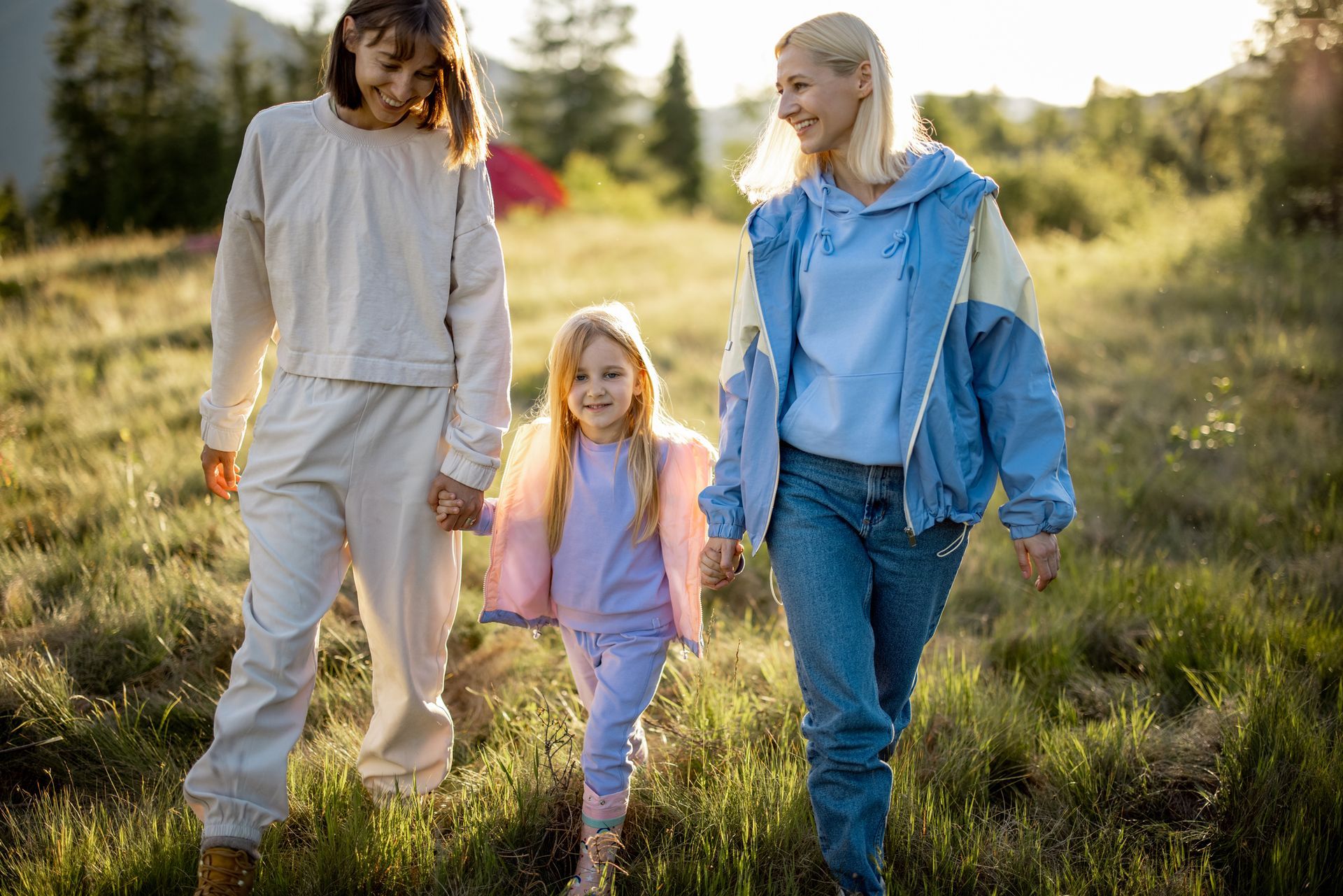 Three children walking through a sunlit grassy field, smiling and holding hands