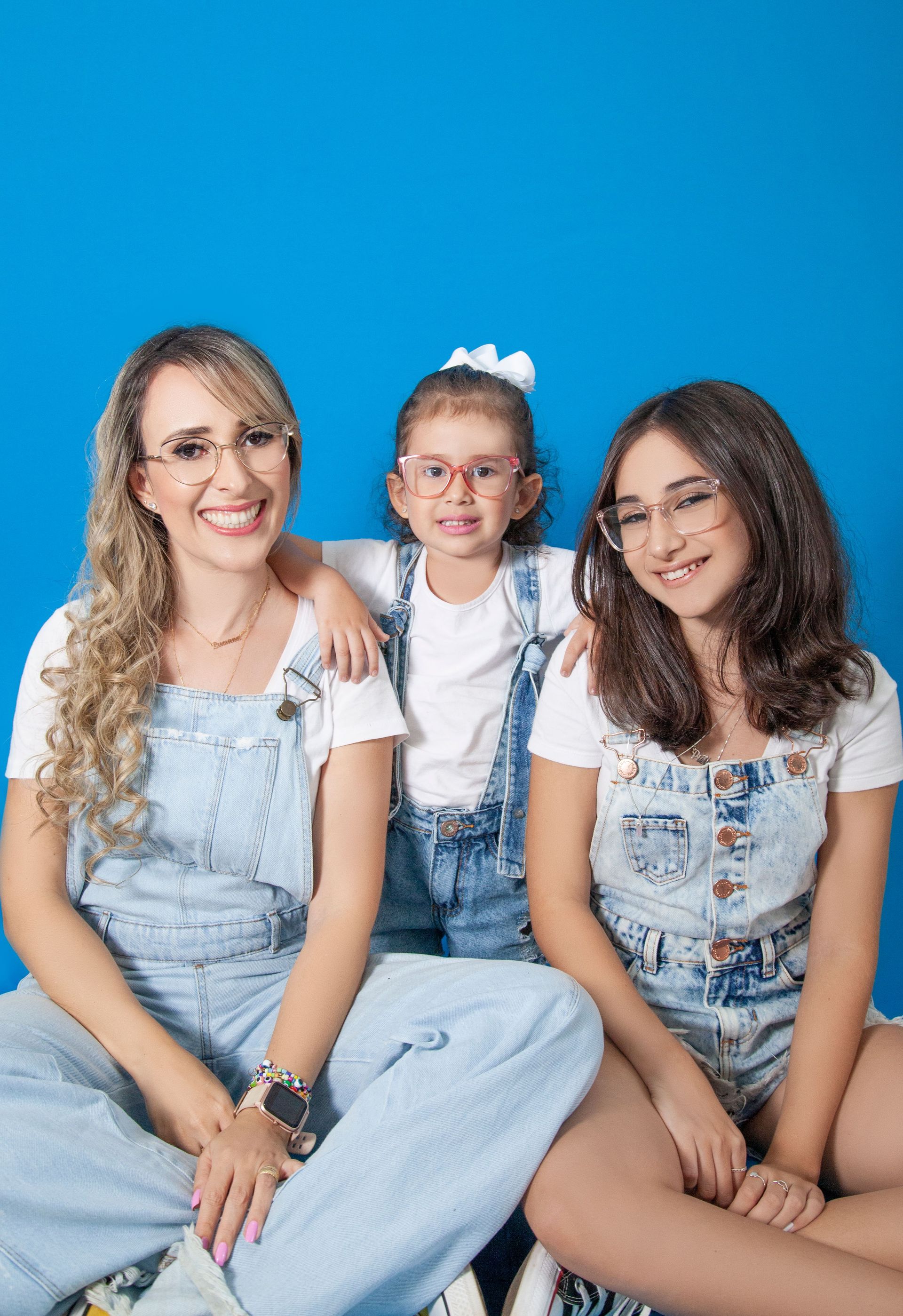 Three smiling people seated against a bright blue background, wearing white tops and denim overalls.