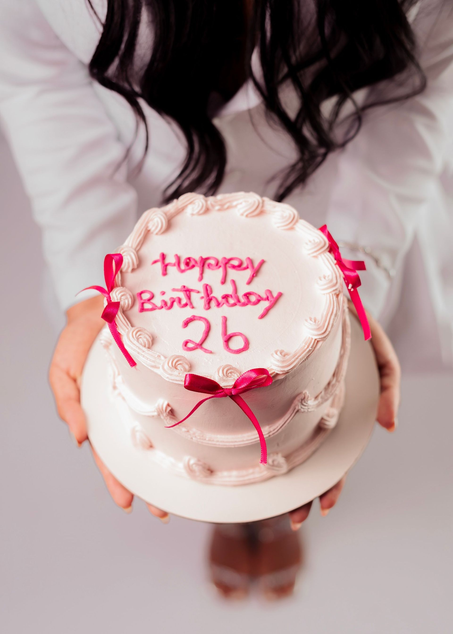Birthday cake with pink ribbons and “Happy Birthday 26” written on top, held by a person