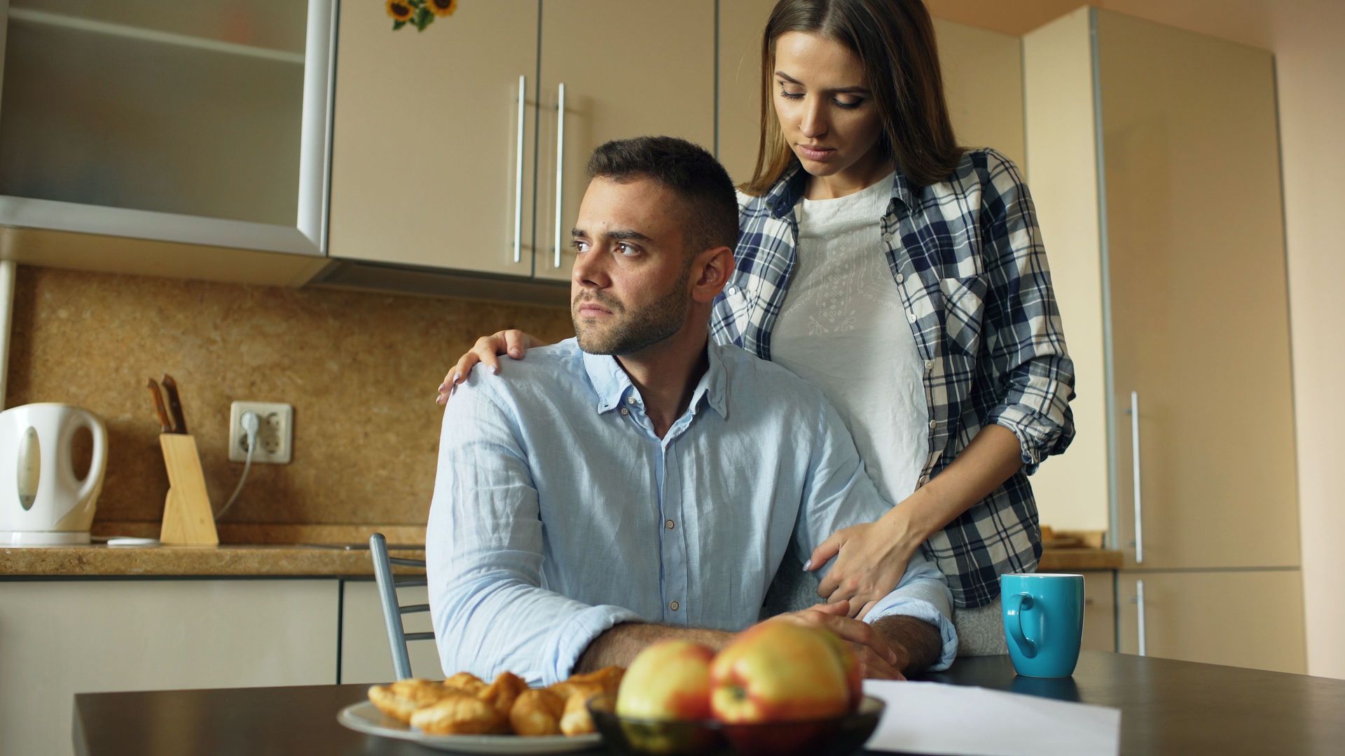 Man and woman in a kitchen, standing by a counter with fruit and mugs.